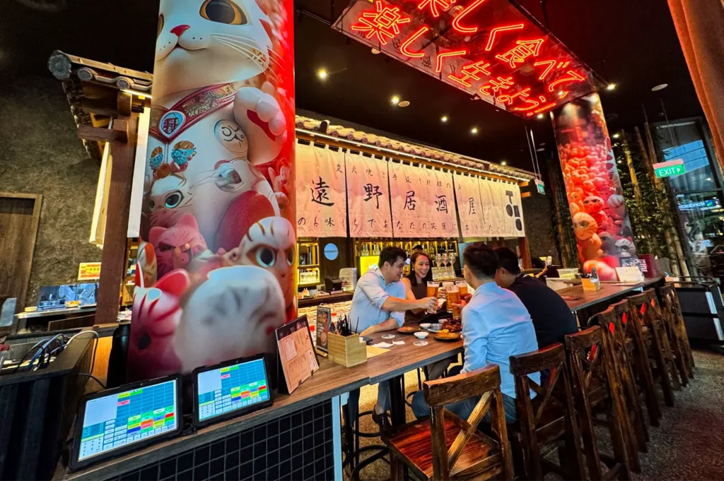 A wide, vibrant eye-level shot captures a group of friends dining inside a lively Japanese restaurant, representing the file "2-Tono Izakaya Review.webp". Four people sit along a long wooden counter-style table, raising their glasses of beer in a celebratory toast under the warm interior lighting. The space is heavily decorated with traditional and modern Japanese elements, including a large pillar wrapped in a colorful graphic of lucky cats (maneki-neko) on the left, a traditional fabric banner (noren) with Japanese calligraphy hanging over the bar area, and a bright red neon sign glowing overhead with Japanese characters. In the foreground on the bottom left, two digital point-of-sale monitor screens display colorful menus, while the background shows a hint of a modern building exterior through a glass pane with a green exit sign.