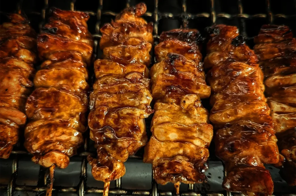 An extreme close-up, top-down shot captures a row of yakitori skewers cooking side-by-side on a wire grill grate, representing the file "2-Types of Yakitori Chicken Skewers in Singapore.webp". Each bamboo skewer is tightly threaded with glistening pieces of chicken, completely coated in a thick, savory dark tare sauce that has caramelized with the heat. The meat shows tempting golden-brown textures and slight charring along the edges from the open flame. The dark background beneath the wire mesh keeps the visual focus entirely on the rich color and succulent appearance of the grilling meat.