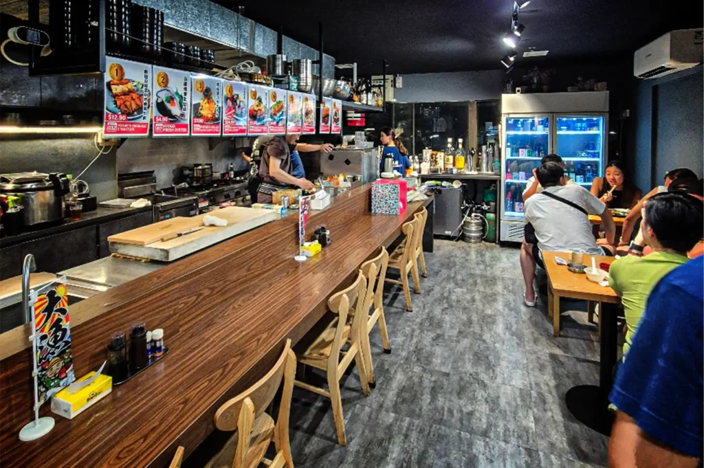 An eye-level, wide shot captures the lively interior of a contemporary Japanese eatery, representing the file "3-Best Fortune Centre Food for Japanese Dining.webp". A long, polished wood-grain counter with light-colored wooden chairs stretches along the left side of the gray, stone-textured floor, separating the dining area from an open kitchen where chefs in dark uniforms are actively preparing meals. Above the kitchen counter, a row of colorful, numbered menu posters with food illustrations and prices are illuminated by overhead track lighting, while stacked black bowls rest on a shelf above. On the right side of the casual dining room, a group of patrons sit at a wooden table enjoying their meal near a large, glass-door beverage cooler glowing with blue light, all set against a dark ceiling and minimalist dark walls.