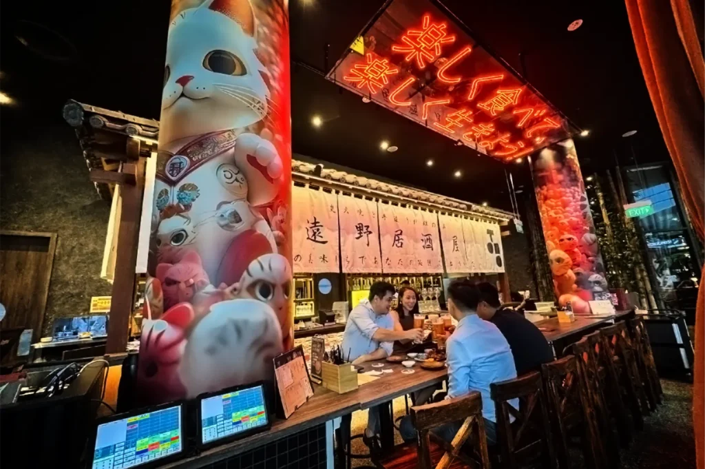 A wide, eye-level shot captures a lively dining group at a vibrant, Japanese-style izakaya counter, representing the file "3-Best Yakitori in Marina Bay Sands Food 6 Spots.webp". Four diners sit on high wooden barstools, raising their glasses in a toast along a long wooden bar topped with small plates of food. The bar is framed by two massive, brightly lit cylindrical pillars wrapped in colorful graphics of beckoning lucky cats (maneki-neko) and various whimsical feline characters. Above the bar counter, traditional white fabric noren curtains printed with Japanese kanji hang below a tiled awning, while a large, glowing red neon sign with Japanese characters stretches across the dark ceiling, creating a bustling, energetic, and contemporary nightlife atmosphere.
