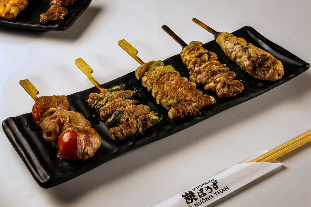 A high-angle shot of a long, wavy black ceramic plate presenting five different Japanese skewers arranged neatly in a row on a white surface. From left to right, the selection features cherry tomatoes wrapped in sliced pork or bacon, meat skewers grilled with green vegetables, and a golden-brown minced chicken tsukune skewer on the far right. A white paper chopstick sleeve with black Japanese and Vietnamese text sits in the bottom right corner.