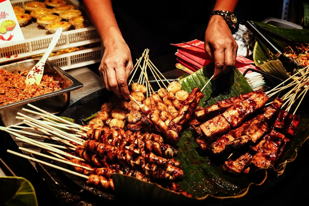 A close-up, eye-level shot captures a vendor's hands expertly arranging a variety of grilled skewers on large, vibrant green banana leaves at a bustling street food market. The assortment includes glistening, dark-glazed chicken or meat spirals, charred tofu blocks, and round fish balls or scallops, all heavily coated in a rich, savory sauce. In the background, stainless steel containers hold spicy condiments and toppings, while a white plastic crate is filled with golden-brown egg tart pastries, creating a warm and appetizing atmosphere under bright, focused market lighting.