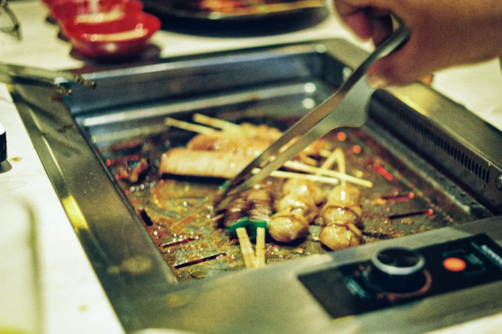 A close-up, eye-level shot captures a person using metal tongs to flip various skewers cooking on a tabletop electric grill. The grill surface is filled with an assortment of food on bamboo sticks, including round meat or fish balls, sliced meat, and vegetables, all sizzling over radiant heat. The stainless steel grill unit features a control panel with a prominent dial in the bottom right corner, while the background is softly blurred, showing red dipping bowls and other dining accessories on the table.