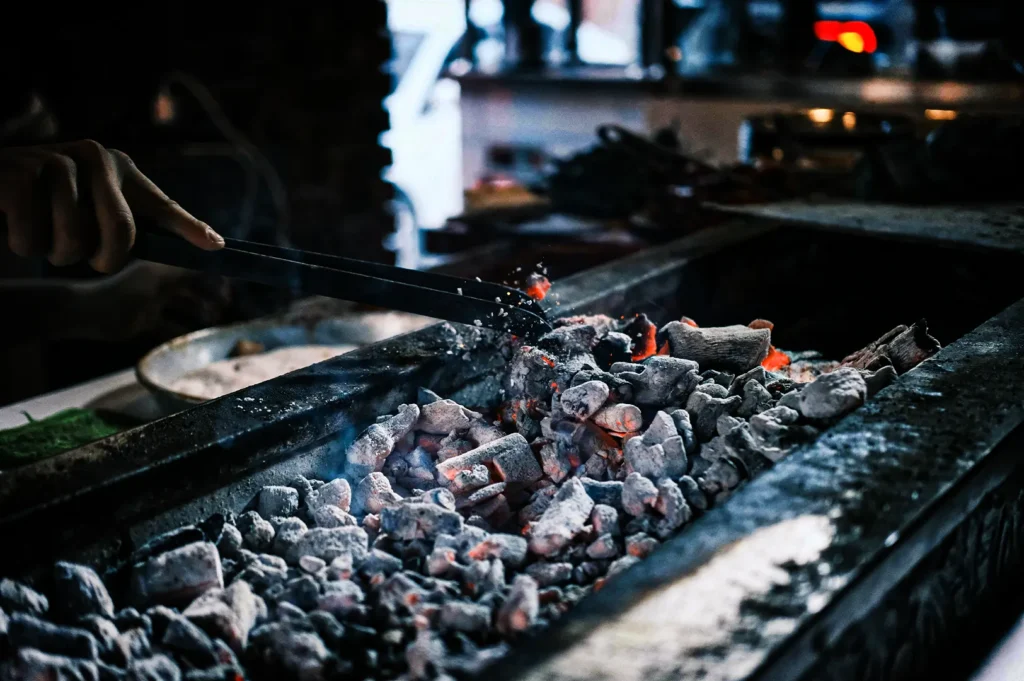 A close-up shot captures a chef's hand using long metal tongs to adjust a bed of glowing binchotan charcoal inside a narrow, rectangular grill station. The charcoal pieces are coated in white ash, with bright red and orange embers burning intensely underneath, sending up faint wisps of smoke and tiny sparks. The background is dimly lit and softly blurred, emphasizing the intense heat and textures of the active grilling setup.