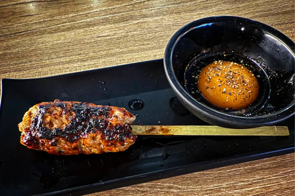 A high-angle, close-up shot captures a single Japanese tsukune skewer served on a sleek, black rectangular plate, representing the file "3-TORIMARO Singapore Review.webp". On the left side of the plate sits a thick, oblong minced chicken meatball molded onto a flat wooden paddle skewer, its surface boasting a glistening, caramelized glaze and prominent dark charred grill marks. To its right, a small, round black ceramic bowl holds a vibrant, glossy raw egg yolk resting in a shallow pool of dark sauce and heavily garnished with coarsely cracked black pepper. The entire plate rests on a light-colored wood-grain table, with overhead lighting creating soft reflections on the plate and highlighting the rich textures of this classic izakaya dish.