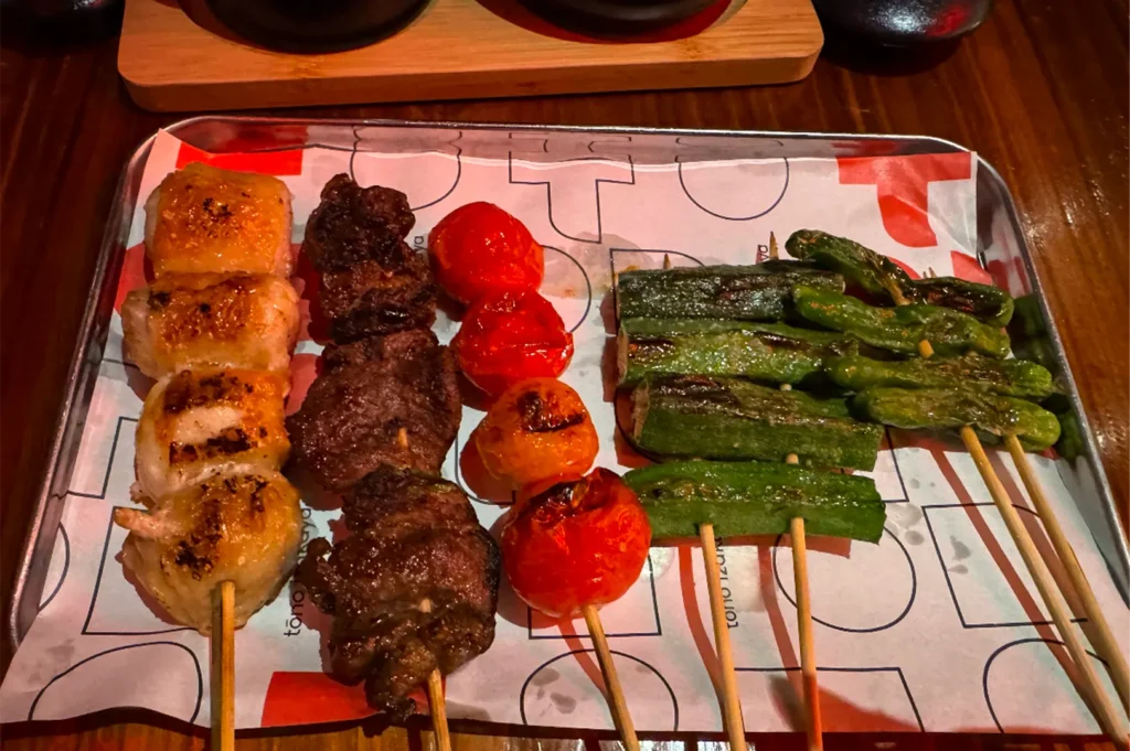 A high-angle, close-up shot displays an assortment of Japanese kushiyaki and yakitori skewers neatly arranged side-by-side on a metal tray, representing the file "3-Tono Izakaya Review.webp". From left to right, the five bamboo skewers hold thick, golden-brown grilled chicken pieces, deeply browned chunks of seasoned meat, shiny whole grilled cherry tomatoes with light charring, cut pieces of bright green okra, and slender green shishito peppers. The skewers rest on a white liner paper printed with red and black geometric designs and text, which is set inside the metal serving tray on a dark wooden table under warm restaurant lighting.