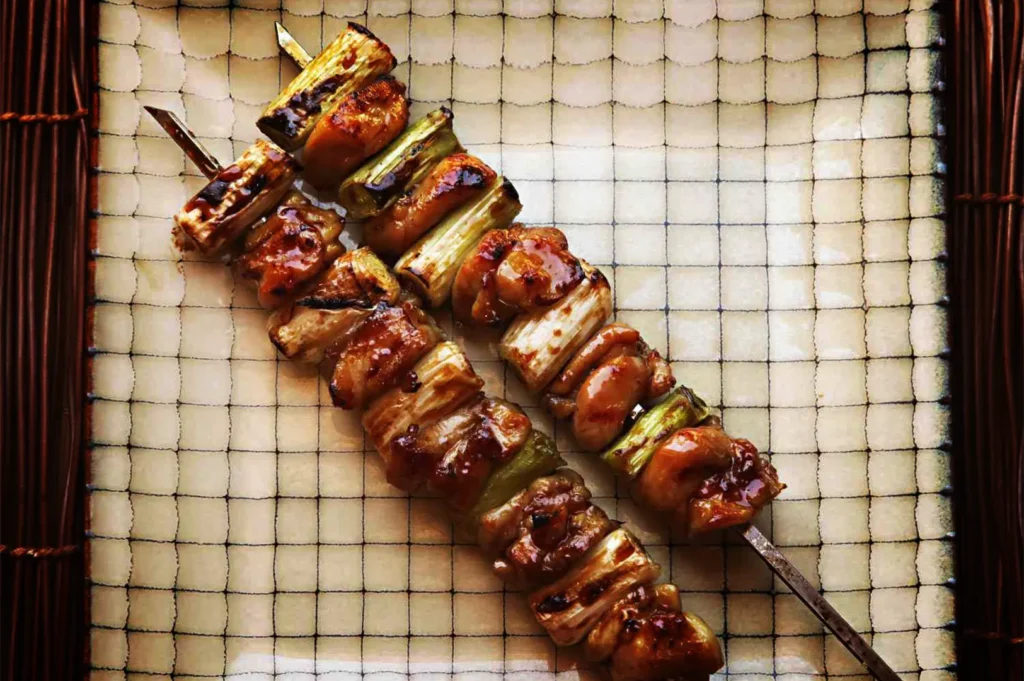 A top-down, overhead shot features two negima yakitori skewers arranged diagonally across a square ceramic plate, representing the file "3-Types of Yakitori Chicken Skewers in Singapore.webp". Each metal skewer alternates between juicy, grilled pieces of chicken thigh coated in a glossy, savory tare sauce and thick chunks of leek that show appetizing char marks from grilling. The plate has a distinct cream color with a subtle, dark grid-like pattern, and it rests on a textured dark wood or bamboo placemat visible on the left and right edges, creating a clean and appetizing presentation.