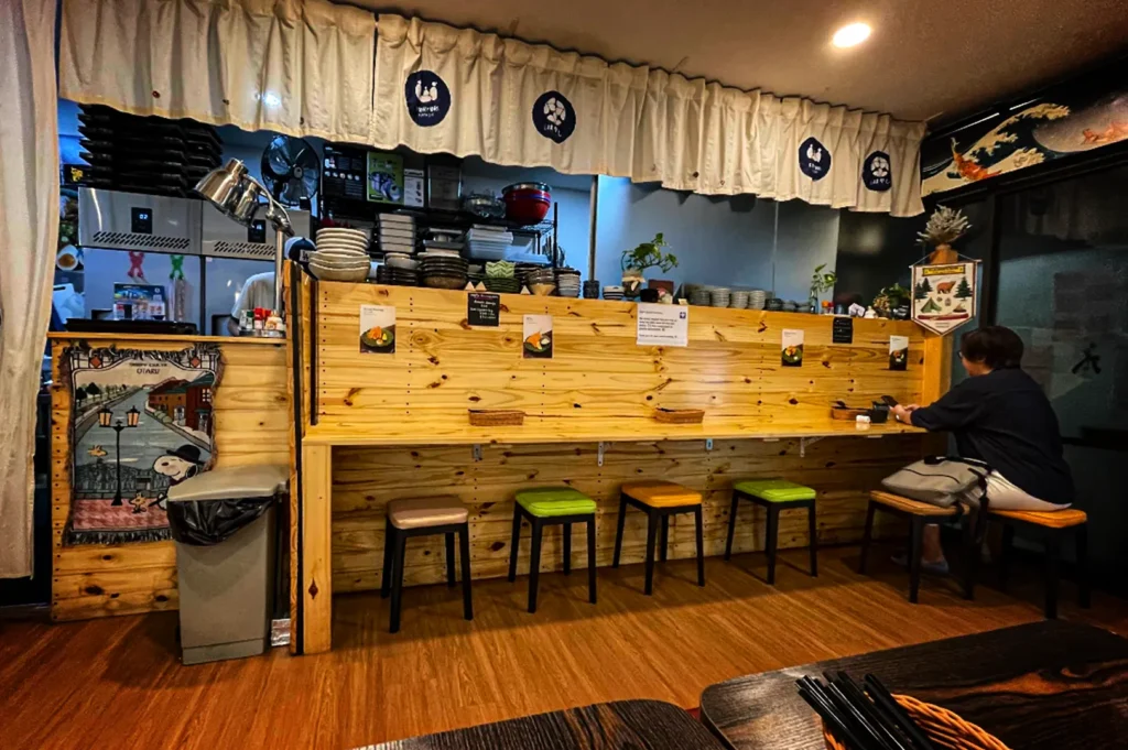 An eye-level, medium shot captures the charming and casual interior of a Japanese restaurant counter area, representing the file "4-Best Fortune Centre Food for Japanese Dining.webp". A long, light-colored knotty pine wooden counter partition runs horizontally across the frame, lined with simple backless stools featuring multi-colored seats in beige, green, and orange over a polished hardwood floor. On the far right, a lone diner sits at the counter looking at their phone, while the wooden partition wall itself is decorated with small flyers, menus, and woven baskets for utensils. Above the partition, a white frilled valence curtain featuring blue circular Japanese crests partially obscures a busy kitchen area filled with stacked plates, bowls, and equipment, creating a cozy and welcoming neighborhood dining atmosphere.