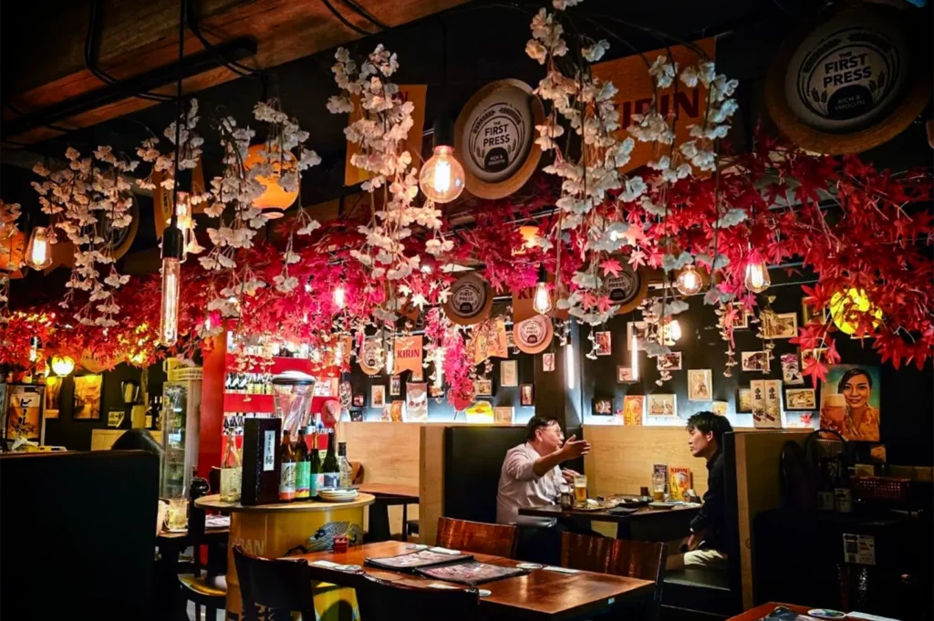 A medium, eye-level shot captures the intricately decorated, cozy interior of a Japanese bar or izakaya, representing the file "4-Best Yakitori in Marina Bay Sands Food 6 Spots.webp". Two men sit in a wooden booth in the center, engaged in conversation over drinks and small plates, while empty wooden dining tables and chairs occupy the foreground. The high, dark ceiling is densely hung with a vibrant canopy of artificial white cherry blossoms and red maple leaves, interwoven with warm, glowing Edison bulb pendant lights. The back wall is adorned with numerous small framed pictures, Kirin beer advertisements, and circular "The First Press" signs, while a yellow barrel stacked with sake bottles stands to the left, creating a lively, authentic, and atmospheric dining environment.