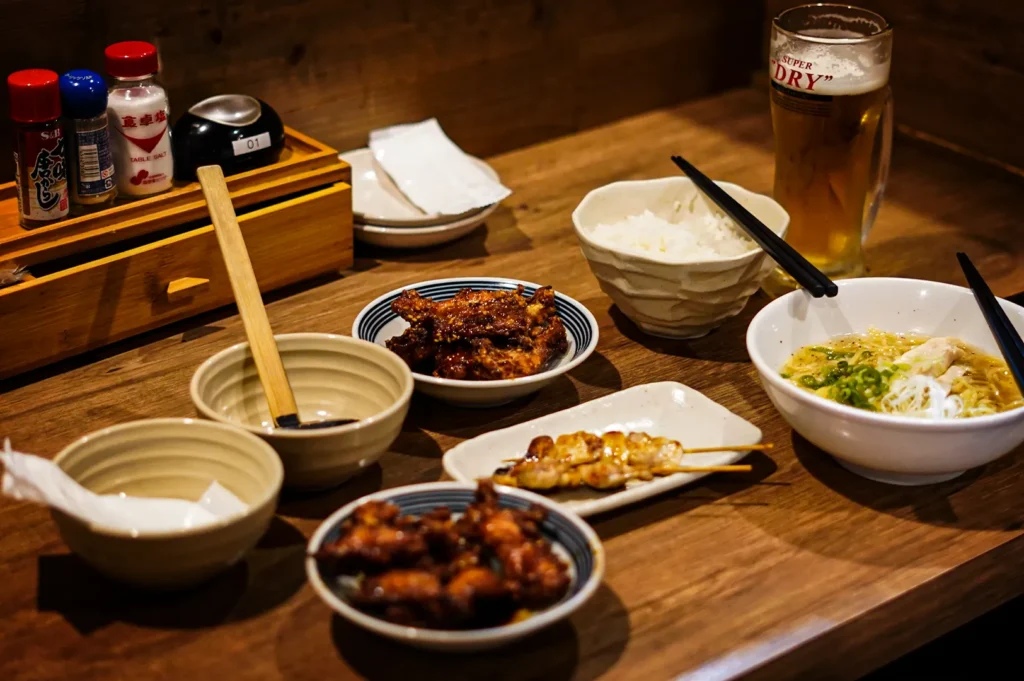 A close-up shot focusing on a long, narrow white plate holding four grilled chicken yakitori skewers, lightly glazed and resting on a wooden table. In the background, a person's hands are blurred as they use chopsticks to lift a piece of meat from another dish. A small ceramic bowl containing a side sauce sits nearby on the left.