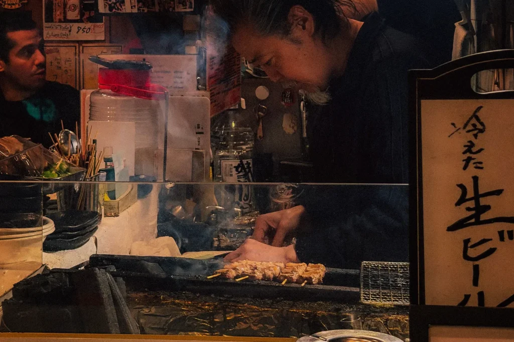 An eye-level shot captures a chef intensely focused on grilling yakitori skewers inside a small, atmospheric Japanese eatery, representing the file "4-Not Just a Stick The Role of Bamboo.jpg". Seen through a clear glass partition that separates the kitchen from the customers, the chef, with grey-streaked hair tied back, leans over a narrow charcoal grill where a row of meat skewers is cooking over hot coals, sending up wisps of smoke. The background is packed with traditional bar details, including stacked black plates, bundled bamboo skewers, and sake bottles, while a customer on the far left looks on from the dimly lit seating area. In the foreground on the right, a vertical wooden sign features hand-painted Japanese calligraphy, completing the authentic, cozy izakaya setting.