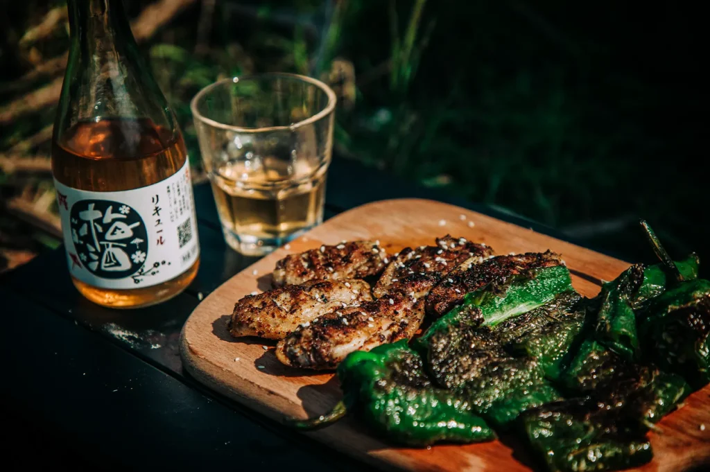 An outdoor, high-angle shot captures a wooden serving board laden with grilled Japanese izakaya dishes, featuring seasoned, sesame-seed-sprinkled chicken wings alongside charred, vibrant green shishito peppers. In the upper-left corner sits an open bottle of Japanese umeshu (plum liqueur) with a white, stylized label, accompanied by a small glass filled with the golden liquid. The setting is bathed in natural sunlight, casting deep shadows across the black table against a soft, dark green foliage background.