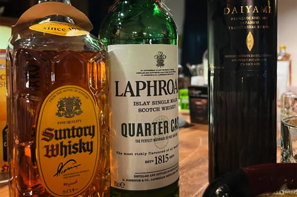 A detailed, close-up shot captures three distinct liquor bottles standing side by side on a smooth wooden counter or table, representing the file "4-TORIMARO Singapore Review.webp". On the left, a textured, tortoise-shell patterned glass bottle of amber-colored Suntory Whisky features a prominent yellow-and-black oval label with a cursive signature. In the center stands a green glass bottle of Laphroaig Islay Single Malt Scotch Whisky featuring a stark white label that reads "Quarter Cask" and displays its establishment date of 1815. To the right, a sleek, dark black bottle of Daiyame Premium Shochu stands tall, highlighting its gold font and traditional Japanese spirit labeling. The tight framing focuses heavily on the labels and bottle designs, while a warm, ambient background softly blurs out the surrounding bar environment.