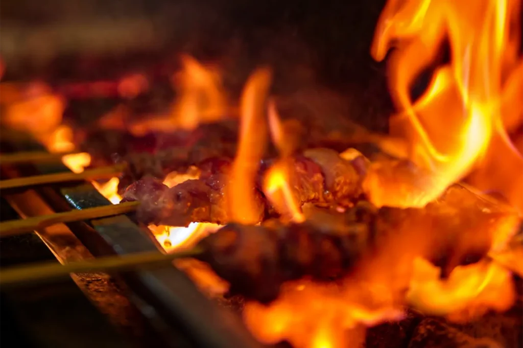 A dramatic, close-up, and low-angle shot captures yakitori skewers cooking over an intense open flame, representing the file "4-Tono Izakaya Review.webp". Several bamboo skewers lined with pieces of meat rest across a metal grill grate, enveloped by vibrant orange and yellow flames that flare up from the hot charcoal below. The intense fire is sharply focused on the right side of the frame, partially obscuring the cooking meat in a warm, bright glow while creating a dramatic, fiery atmosphere that highlights the traditional Japanese grilling process.