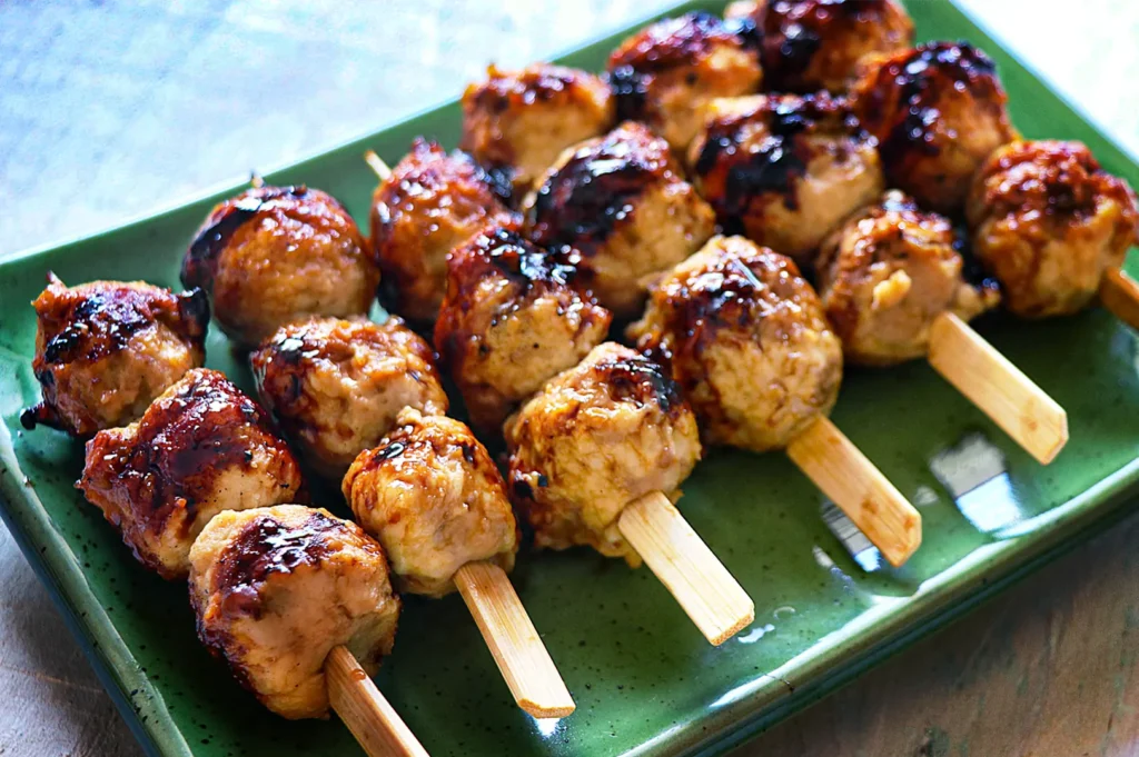 A close-up, high-angle shot captures a platter of tsukune (Japanese chicken meatball) skewers, representing the file "4-Types of Yakitori Chicken Skewers in Singapore.webp". Arranged neatly on a rectangular green glazed ceramic plate, several flat paddle-style bamboo skewers each hold three plump, rounded chicken meatballs. The meatballs feature a golden-brown, textured surface brushed with a glossy, dark tare sauce that shows areas of deep caramelization and appealing dark char marks from being grilled. The vibrant green plate rests on a neutral-toned wooden surface, emphasizing the savory and succulent appearance of the dish.