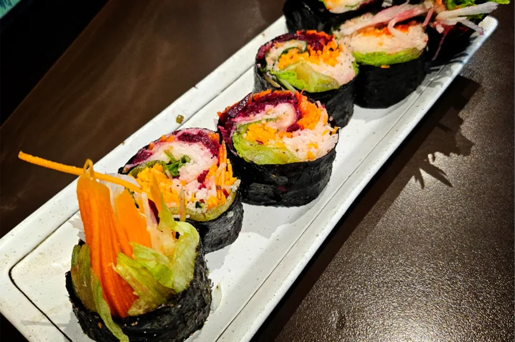 A high-angle, close-up shot captures a vibrant platter of vegetarian sushi rolls arranged on a long, narrow white ceramic dish, representing the file "5-Best Fortune Centre Food for Japanese Dining.webp". The sushi pieces are lined up diagonally across the frame, wrapped tightly in dark nori seaweed and packed with a colorful assortment of fresh fillings, including shredded carrots, green lettuce, white radish, and deep purple beetroot. The piece in the bottom-left foreground stands out as an end-roll, featuring dramatic, tall tufts of julienned orange carrots and crisp green greens bursting out of the top. The entire dish rests on a dark, textured brown tabletop, with focused overhead lighting casting subtle shadows that emphasize the fresh and healthy textures of the meal.