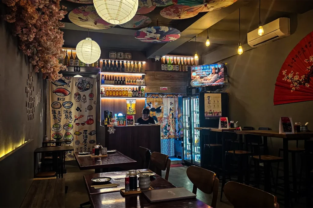 A wide, eye-level shot captures the intimate, atmospheric interior of a Japanese izakaya, representing the file "5-Best Yakitori in Marina Bay Sands Food 6 Spots.webp". In the center background, a staff member stands behind a dark wooden service counter backed by shelves neatly stacked with illuminated sake and liquor bottles, flanked by colorful fabric curtains displaying traditional Japanese illustrations and geisha motifs. The dining area is furnished with dark wood tables and chairs in the foreground and a row of high-top bar tables on the right. The ceiling is intricately decorated with a canopy of patterned paper umbrellas and large, glowing white paper lanterns, complemented by warm, hanging Edison bulbs and a bunch of pink cherry blossoms on the left wall, creating a cozy and culturally rich dining ambiance.