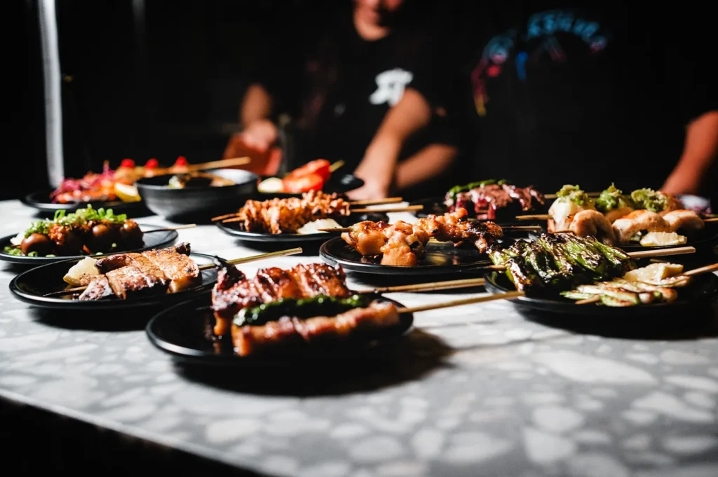 A low-angle, shallow depth-of-field shot of a stone counter laden with numerous small black plates of Japanese izakaya dishes. In the foreground, grilled skewers including pork belly and charred green peppers are in sharp focus. The middle and background feature an assortment of other skewers, such as chicken yakitori, shiitake mushrooms, and sliced tomatoes, stretching across the counter. In the softly blurred background, chefs in black uniforms work behind the line.