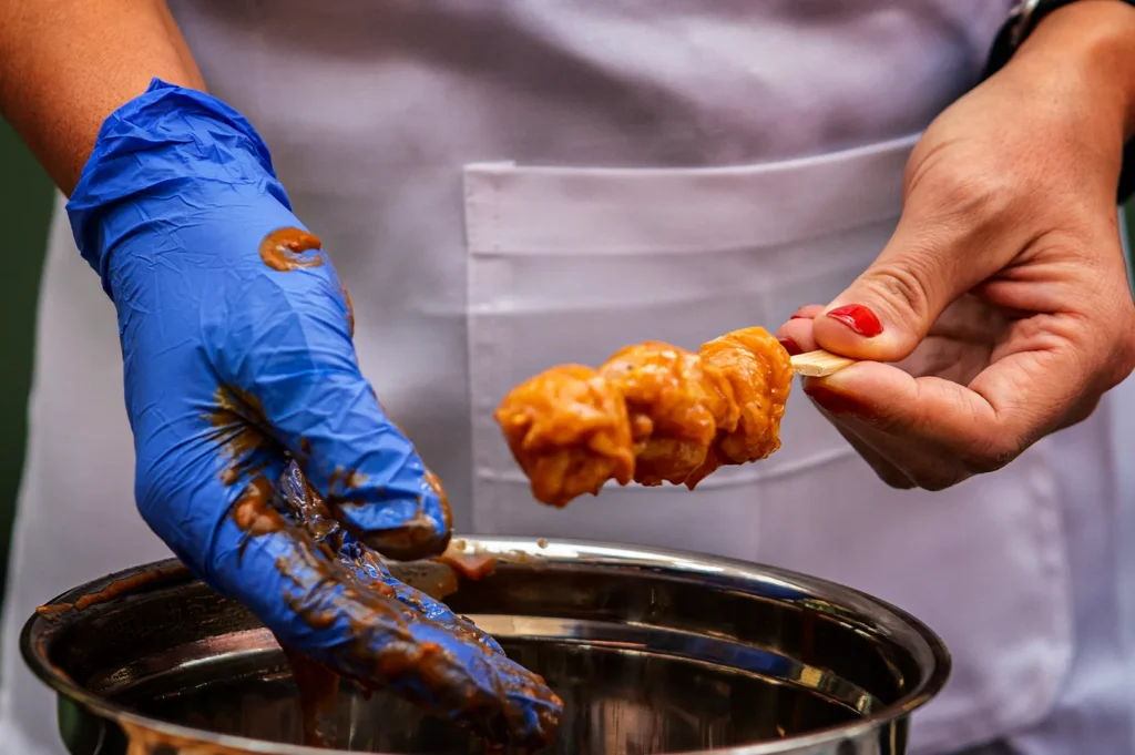 A close-up shot captures the preparation of a street food skewer, representing the file "5-Not Just a Stick The Role of Bamboo.webp". A person wearing a white apron holds a flat bamboo paddle skewer with one hand, featuring red painted fingernails, showcasing three round food pieces completely coated in a thick, orange-brown peanut sauce. The other hand, clad in a bright blue nitrile glove heavily smeared with the same savory sauce, hovers over a stainless steel mixing bowl used to coat the food. The tight focus captures the details of the culinary process, highlighting the preparation stage before cooking or serving.
