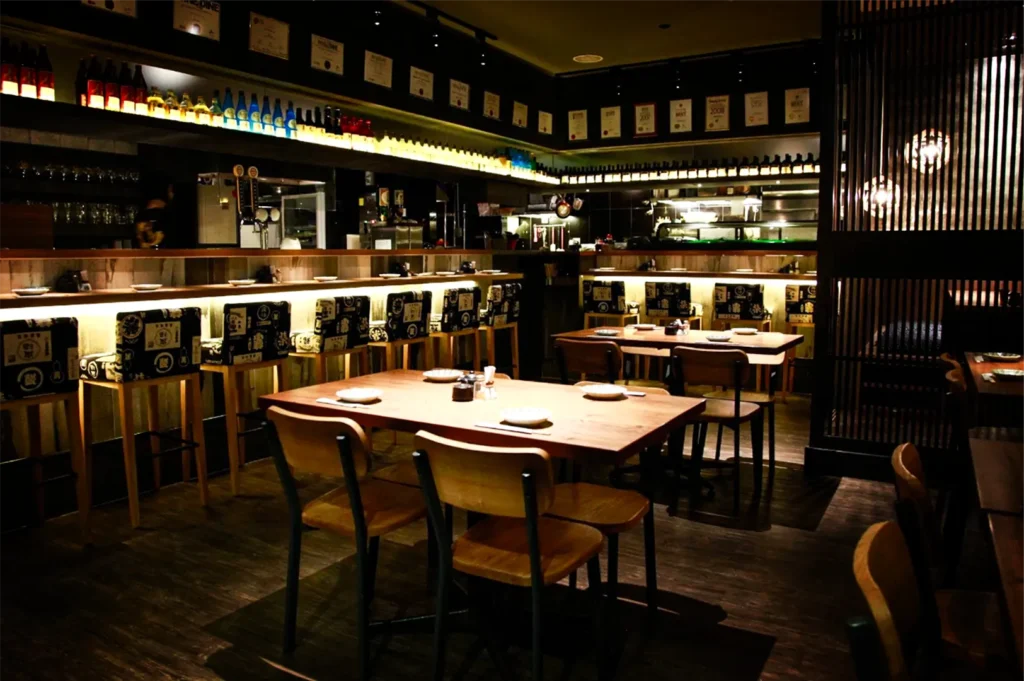A wide, eye-level shot captures the moody, dimly lit interior of a Japanese izakaya restaurant featuring dark wooden floors and furniture. In the foreground, a polished wooden dining table is neatly set with small white plates and chopsticks, surrounded by simple wooden chairs. Behind it, a long, warmly illuminated counter bar is lined with tall barstools upholstered in patterned black-and-white fabric, while rows of sake bottles and framed certificates decorate the upper shelves along the dark walls.