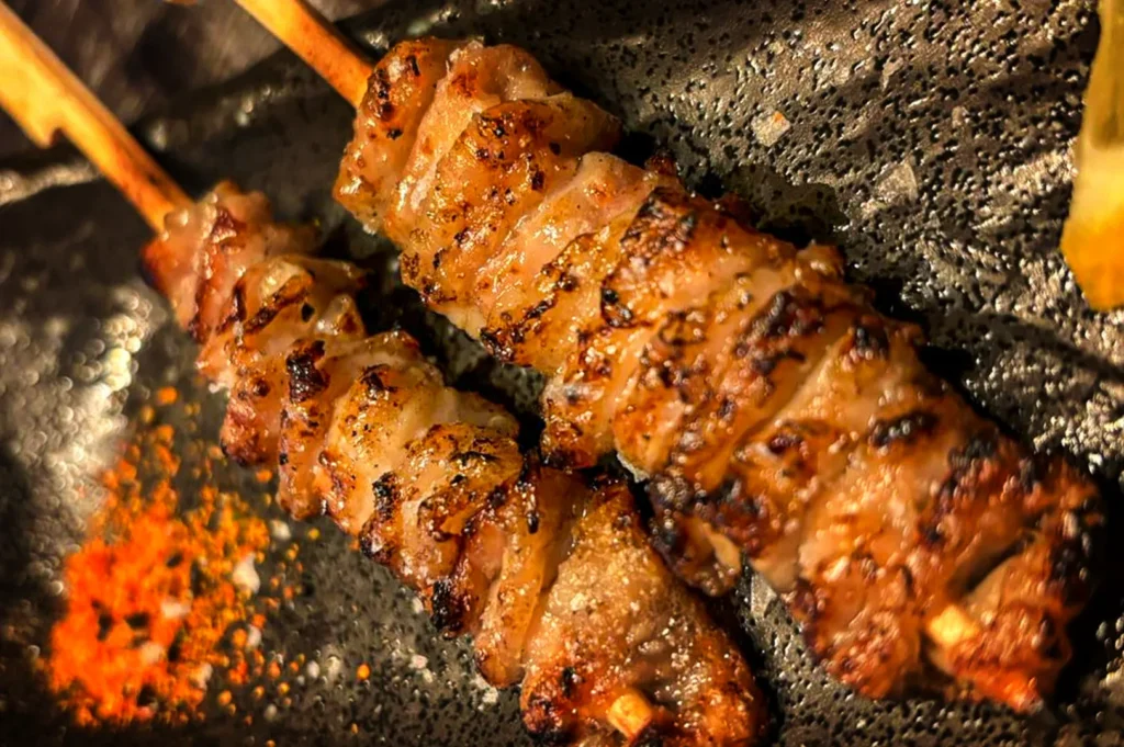 An overhead, close-up shot captures two kawa (chicken skin) yakitori skewers served on a textured surface, representing the file "5-Types of Yakitori Chicken Skewers in Singapore.webp". The bamboo skewers are tightly threaded with folded ribbons of chicken skin that have been grilled to a golden brown, showcasing a glistening, crispy texture with distinct dark char marks along the edges. Placed diagonally on a dark, textured black plate, the skewers are accompanied by a small sprinkle of bright orange-red shichimi togarashi (Japanese seven-spice powder) in the bottom-left corner and a sliver of lemon visible on the far right edge, creating a visually rich and savory presentation.