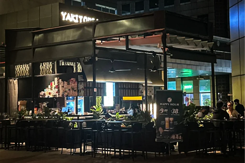 A wide, eye-level shot captures the exterior of an open-air Japanese restaurant at night, representing the file "6-Best Yakitori in Marina Bay Sands Food 6 Spots.webp". The eatery features a dark, modern industrial canopy with a retractable roof and ceiling fans covering a sleek bar and outdoor dining area. On the left, the bar counter is backlit with cool light, displaying a tiered wall of sake labels, while potted green plants line the perimeter. To the right, a prominent "HAPPY HOUR" menu board stands near several diners seated at dark high-top tables, with a glowing "YAKITORI" sign visible on the building structure above, creating a relaxed, urban nightlife vibe.