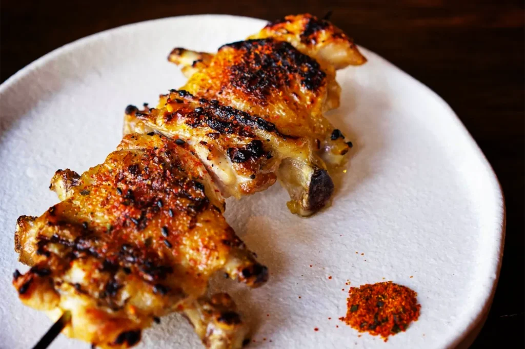 A high-angle, close-up shot captures a teba (chicken wing) yakitori skewer served on a plate, representing the file "6-Types of Yakitori Chicken Skewers in Singapore.webp". The single skewer features two bone-in chicken wing sections grilled to a crispy, golden brown, showcasing heavily charred skin and a generous dusting of red-and-black shichimi togarashi (Japanese seven-spice powder) across the surface. Placed diagonally on a round, textured white ceramic plate, the skewer is accompanied by a small, neat pile of additional spice blend in the lower-right corner, set against a dark, out-of-focus background.