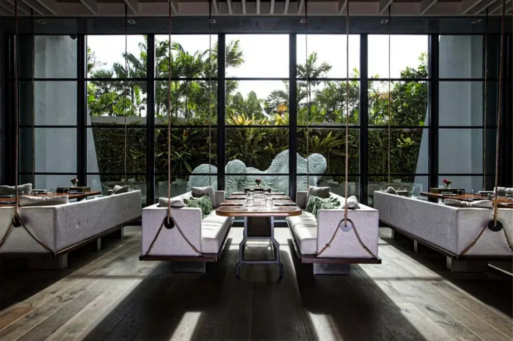 A wide, eye-level shot captures the elegant, modern interior of a luxurious restaurant dining room, representing the file "7-Best Yakitori in Marina Bay Sands Food 6 Spots.webp". Arranged on a dark hardwood floor, the seating features unique, long upholstered grey booths with decorative thick ropes suspended from the ceiling, mimicking indoor swings. The center dining table is set with glasses and utensils, while matching tables flank it on either side. Sunlight streams through a massive, grid-patterned glass window wall that spans the entire background, revealing a lush outdoor garden filled with vibrant green tropical plants, palm trees, and a large, abstract grey wavy sculpture, creating a serene, upscale, and bright dining environment.
