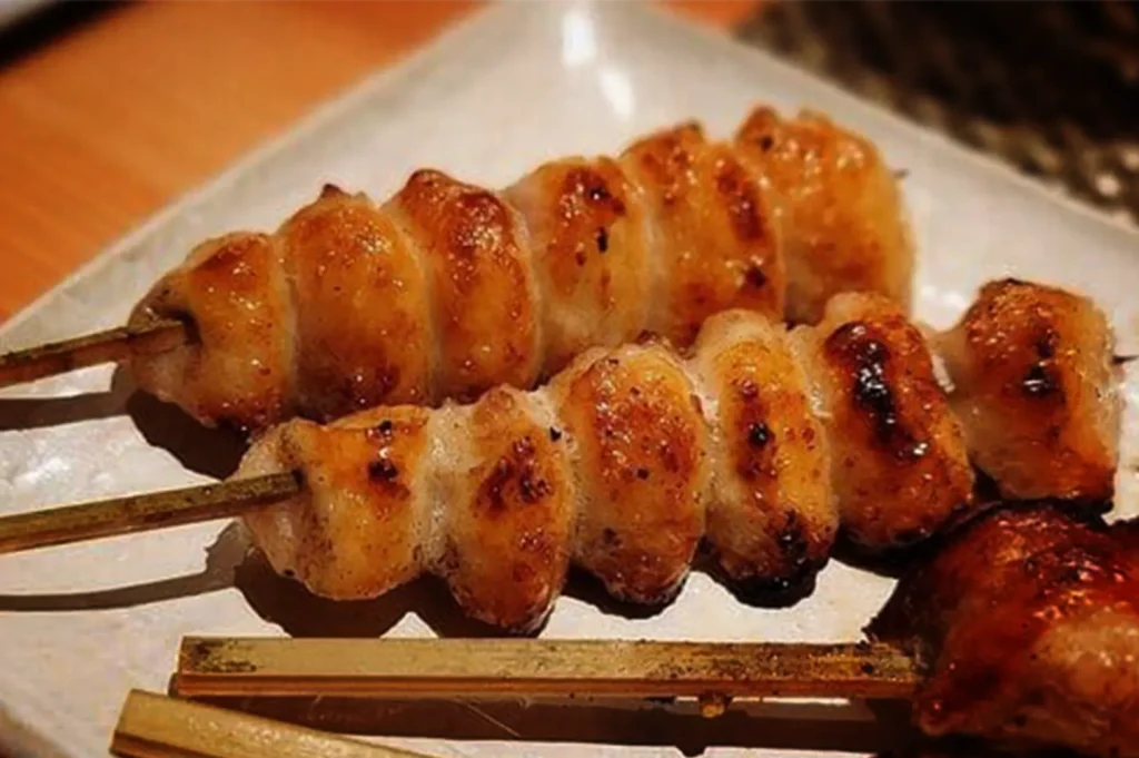A close-up, high-angle shot captures two bonjiri (chicken tail) yakitori skewers served on a platter, representing the file "7-Types of Yakitori Chicken Skewers in Singapore.webp". The bamboo skewers are packed with plump, rounded pieces of grilled chicken tail that have been cooked to a glistening golden-brown finish, displaying crispy edges and small, appetizing char spots. They sit alongside other skewers on a light-colored, textured ceramic rectangular plate, creating a warm and savory presentation set against a softly blurred, neutral background.
