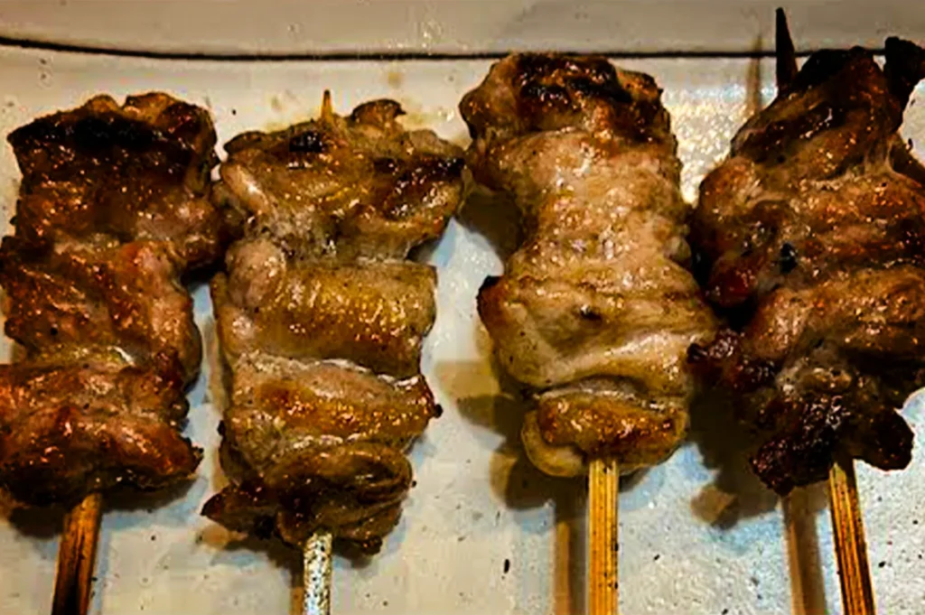 A high-angle, close-up shot captures four grilled yakitori skewers arranged side-by-side on a light-colored rectangular plate. The bamboo and metal skewers are tightly threaded with different cuts of chicken meat, displaying a range of golden-brown textures, glistening juices, and varying degrees of charring from the grill. The skewers on the far left and far right show deeper browning and dark, crispy edges, while the middle skewers feature lighter, succulent folds of meat. The plain, light background emphasizes the rich, savory colors and grilled textures of the seasoned Japanese street food.