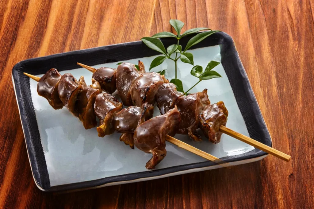 A high-angle, close-up shot captures two hatsu (chicken heart) or sunagimo (gizzard) yakitori skewers arranged diagonally on a serving dish, representing the file "9-Types of Yakitori Chicken Skewers in Singapore.webp". The bamboo skewers hold several thick, plump pieces of organ meat that are generously glazed in a glossy, dark tare sauce, reflecting the light and showing slight charring on the edges from being grilled. They rest on a rectangular ceramic plate with a light grey center and a thick, dark rim, decorated with a small green leafy sprig at the top. The entire plate is set against a warm, rustic wooden table surface with a prominent vertical grain.