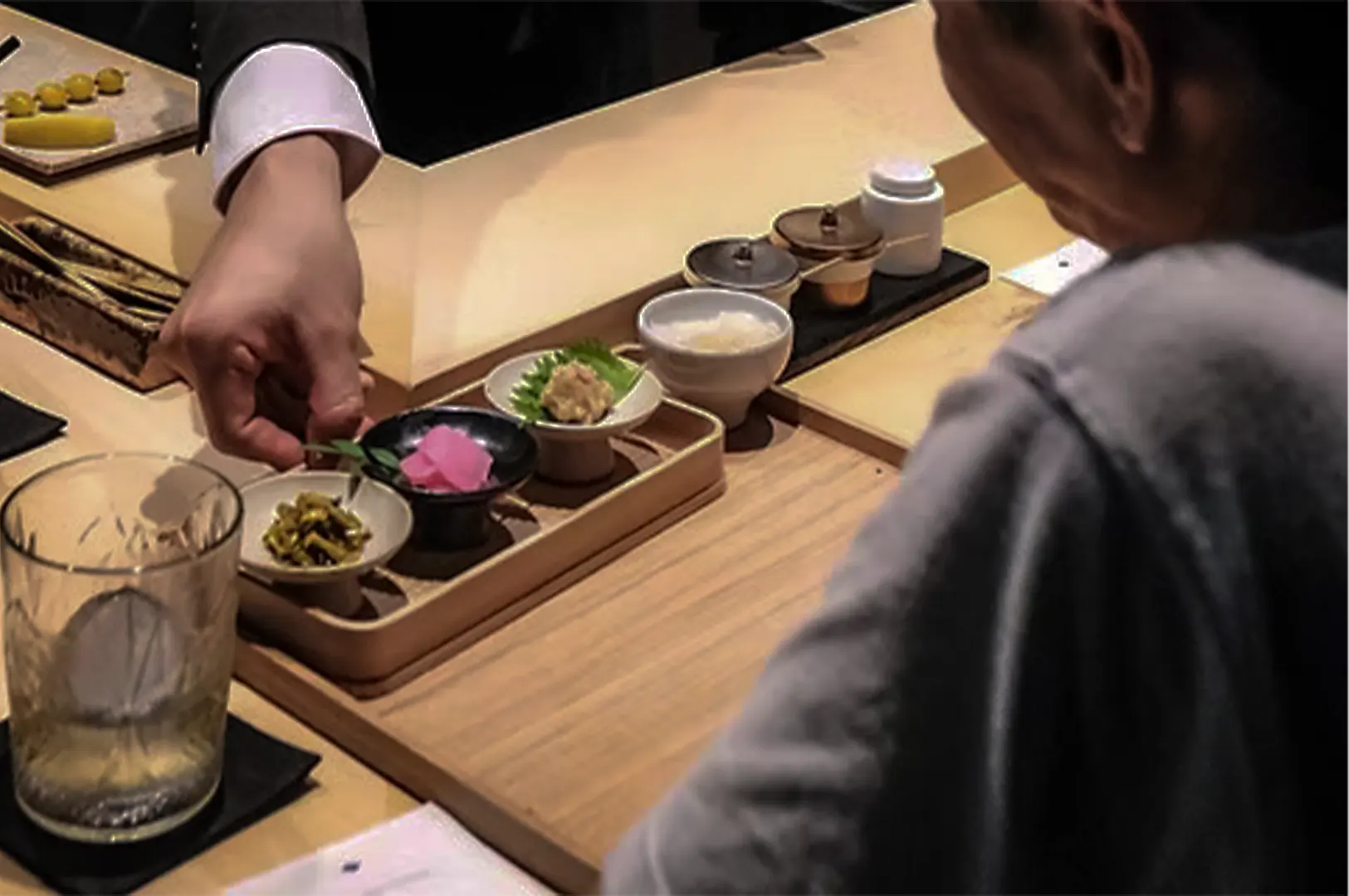 An over-the-shoulder, close-up shot captures a solo diner being served at a wooden counter, representing the file "1-A Table for One The Unexpected Joy of Dining Alone.webp". From a high-angle perspective, a server in a dark suit jacket adjusted with a crisp white cuff carefully places a wooden tray containing three small assorted appetizer dishes—including pickled pink ginger and green garnishes—in front of the customer. A glass of water with a large ice sphere rests in the bottom-left foreground, while small condiment containers line the upper section of the clean, minimalist restaurant bar setup.
