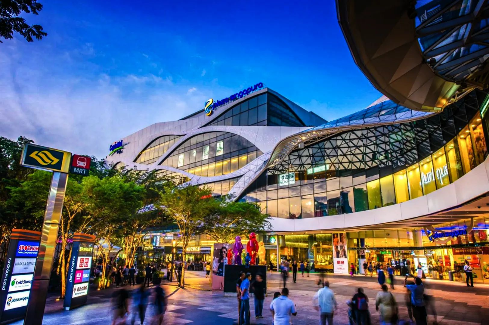 A wide, low-angle shot captures the bustling exterior plaza of a large, modern shopping mall at dusk, representing the file "1-Best Yakitori Near Plaza Singapura.webp". The multi-story building features a striking architectural design with fluid, sweeping white bands and a massive, curved glass facade structured with a geometric web pattern that reflects the deep blue evening sky. On the left, a prominent illuminated yellow and red MRT subway station sign stands amidst a line of green trees and directory boards. Below, a wide pedestrian plaza is filled with blurred figures of people walking by, near brightly lit storefront entrances like Uniqlo, a Starbucks, and a cluster of colorful, glossy bear sculptures standing as a central art installation.