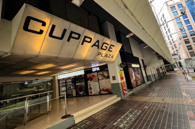 A low-angle, wide shot captures the exterior entrance of a multi-story commercial building walkway, representing the file "1-Best Yakitori in Cuppage Plaza Food Restaurants To Try.webp". A massive, angular backlit sign overhead glows with warm amber light, prominently displaying the words "CUPPAGE PLAZA" in large black block lettering. Below the sign, a set of wide, shallow steps leads into the building's entranceway, where a spa storefront with posters of treatments and a neighboring massage parlor are visible behind glass doors. To the left, a glass railing with a metallic handrail frames a ramp or stairwell leading down, while the right side of the frame features a patterned brick-tiled pedestrian sidewalk stretching into the background along the building's facade.