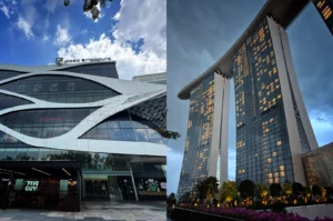 This split-composition image contrasts two prominent architectural landmarks in Singapore, representing the file "1-Plaza Singapura vs Marina Bay Sands.webp". On the left side, a low-angle shot during the day highlights the modern exterior of the Plaza Singapura shopping mall, showcasing its distinctive white, undulating ribbon-like facade accents wrapping around large glass windows, with a "Five Guys" restaurant entrance visible at the street level under a bright blue sky with fluffy white clouds. On the right side, a dramatic low-angle evening shot captures two of the soaring towers of the Marina Bay Sands hotel, illuminated by glowing interior room windows under a moody, overcast twilight sky. The towers are crowned by the iconic boat-shaped SkyPark cantilever structure, while manicured green trees and bright purple flowers line the walkway at the base.