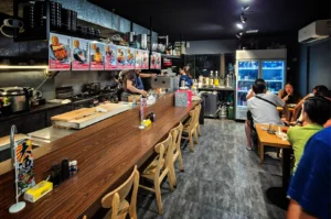 A wide, eye-level shot captures the bright, modern interior of a Japanese restaurant, representing the file "1-Sakedokoro Eizaburo Singapore.webp". On the left, a long, angled wooden bar counter dominates the space, set with empty wooden chairs, condiment bottles, and a large wooden cutting board. Behind the counter, kitchen staff in dark t-shirts work diligently in the open kitchen, which is lined with equipment, hanging black bowls, and a row of colorful menu posters displaying dish photos and prices. To the right, a gray stone-patterned floor leads toward a small dining area where patrons are seated at wooden tables, a large glass-door beverage refrigerator glowing with blue light, and a bar station stocked with liquor bottles in the background.