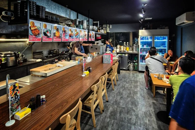 A wide, eye-level shot captures the bright, modern interior of a Japanese restaurant, representing the file "1-Sakedokoro Eizaburo Singapore.webp". On the left, a long, angled wooden bar counter dominates the space, set with empty wooden chairs, condiment bottles, and a large wooden cutting board. Behind the counter, kitchen staff in dark t-shirts work diligently in the open kitchen, which is lined with equipment, hanging black bowls, and a row of colorful menu posters displaying dish photos and prices. To the right, a gray stone-patterned floor leads toward a small dining area where patrons are seated at wooden tables, a large glass-door beverage refrigerator glowing with blue light, and a bar station stocked with liquor bottles in the background.