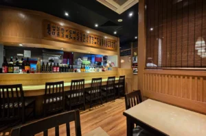 A wide-angle, eye-level indoor shot captures the warm and traditional interior of an izakaya or Japanese restaurant, representing the file "1-Shinjuku Restaurant Cuppage.webp". Centered in the background is a long wooden dining counter lined with several high-backed dark wooden chairs, with an array of sake bottles arranged along its ledge. Above the open kitchen counter, where a chef wearing a white cap can be seen working, hangs a long, decorative wooden menu board featuring vertical Japanese calligraphy characters. The foreground showcases cozy wooden tables and chairs set against polished wooden floors, with a slatted wooden screen partition on the right enhancing the authentic, intimate dining atmosphere under soft spotlighting.