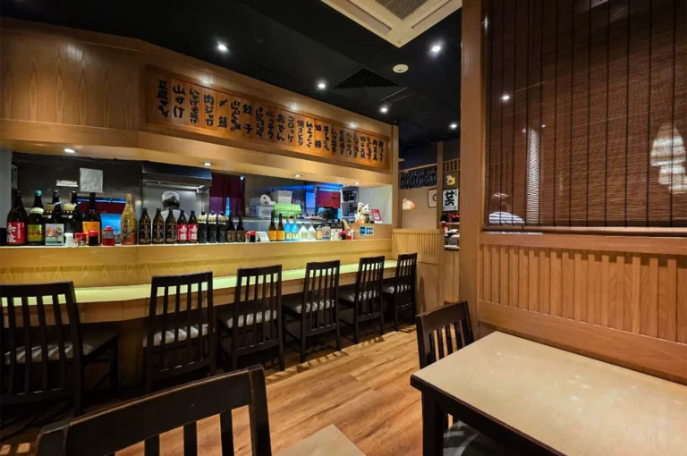 A wide-angle, eye-level indoor shot captures the warm and traditional interior of an izakaya or Japanese restaurant, representing the file "1-Shinjuku Restaurant Cuppage.webp". Centered in the background is a long wooden dining counter lined with several high-backed dark wooden chairs, with an array of sake bottles arranged along its ledge. Above the open kitchen counter, where a chef wearing a white cap can be seen working, hangs a long, decorative wooden menu board featuring vertical Japanese calligraphy characters. The foreground showcases cozy wooden tables and chairs set against polished wooden floors, with a slatted wooden screen partition on the right enhancing the authentic, intimate dining atmosphere under soft spotlighting.