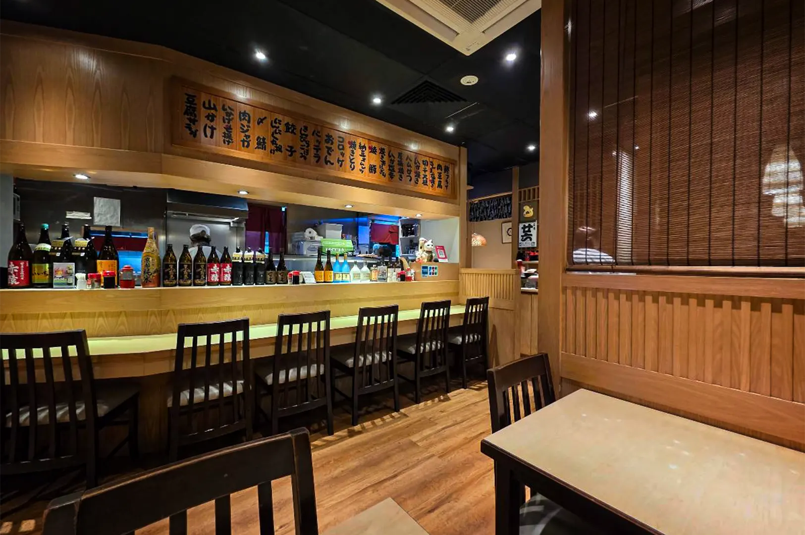 A wide-angle, eye-level indoor shot captures the warm and traditional interior of an izakaya or Japanese restaurant, representing the file "1-Shinjuku Restaurant Cuppage.webp". Centered in the background is a long wooden dining counter lined with several high-backed dark wooden chairs, with an array of sake bottles arranged along its ledge. Above the open kitchen counter, where a chef wearing a white cap can be seen working, hangs a long, decorative wooden menu board featuring vertical Japanese calligraphy characters. The foreground showcases cozy wooden tables and chairs set against polished wooden floors, with a slatted wooden screen partition on the right enhancing the authentic, intimate dining atmosphere under soft spotlighting.