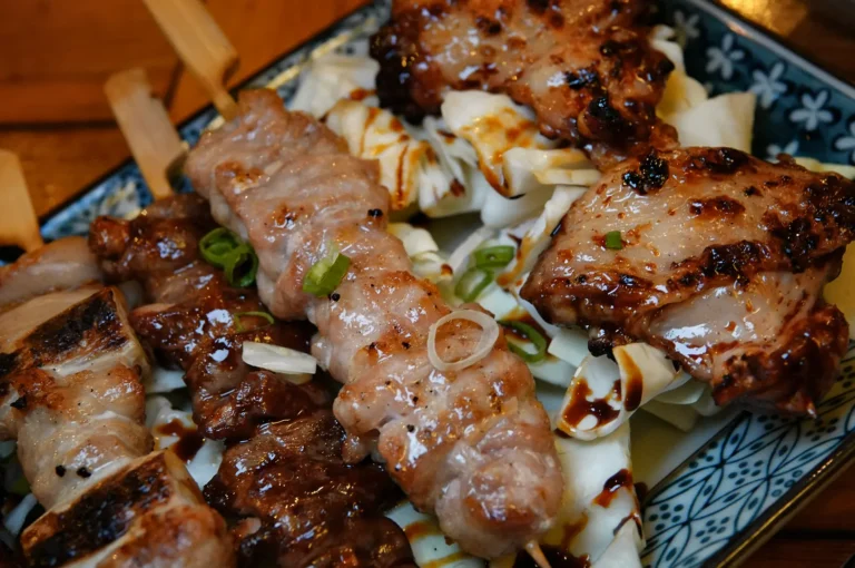 A close-up, high-angle shot captures an assortment of grilled yakitori skewers and meat pieces drizzled with a dark, glossy sauce and garnished with chopped green onions. The skewered meats rest over a bed of raw white cabbage leaves inside a decorated, patterned rectangular plate.