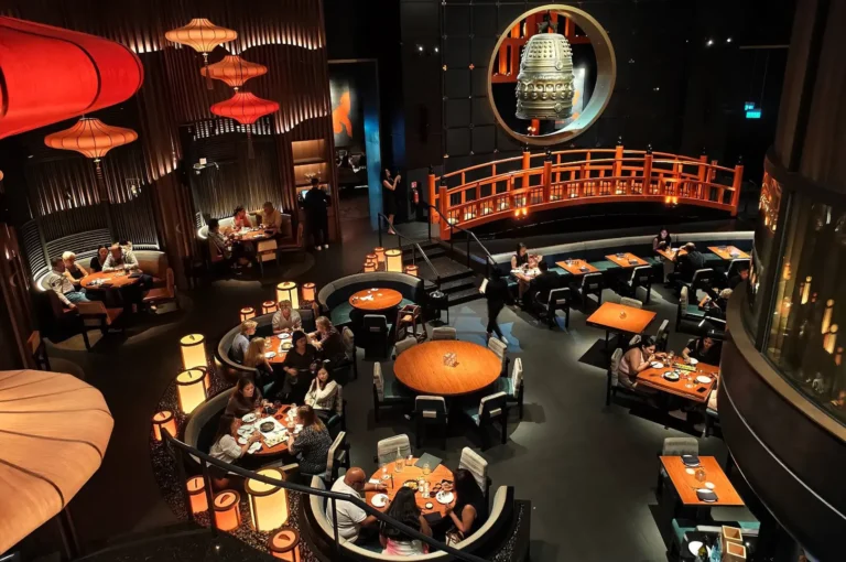 A high-angle, wide shot captures the dramatic, upscale dining room of a multi-level restaurant with a spectacular Japanese-inspired interior design, representing the file "1-The KOMA Experience.webp". Numerous circular and rectangular wooden tables are spread across the main floor, occupied by patrons dining under soft, warm lighting. The striking multi-level space is anchored in the background by an elevated, bright orange traditional footbridge that spans across the back wall, situated just beneath a massive, circular cutout displaying a monumental bronze Japanese temple bell. The sophisticated, theatrical atmosphere is enhanced by a towering wall of slatted wood paneling on the left, flanked by large glowing paper lanterns in orange and cream hues that illuminate the cozy booth seating below.