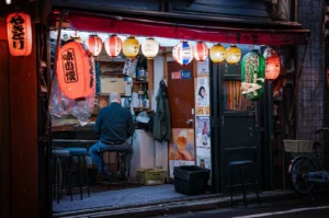 A wide, eye-level shot captures a quiet and intimate nighttime street scene at a traditional Japanese izakaya, representing the file "1-The Last Customer at the Counter Staying Until the End.webp". An elderly, bald man wearing a dark long-sleeve shirt and blue jeans sits on a low stool with his back to the camera, looking toward the interior counter of a small, open-front restaurant. The storefront is framed by a low red awning and illuminated by a bright, vibrant row of hanging paper lanterns in shades of red, white, yellow, and green adorned with Japanese text and logos. Vintage beverage posters and a green coat hang on the wall next to a sliding door, while empty stools, plastic crates, and a bicycle parked on the right emphasize the calm, end-of-the-night atmosphere of the alleyway.