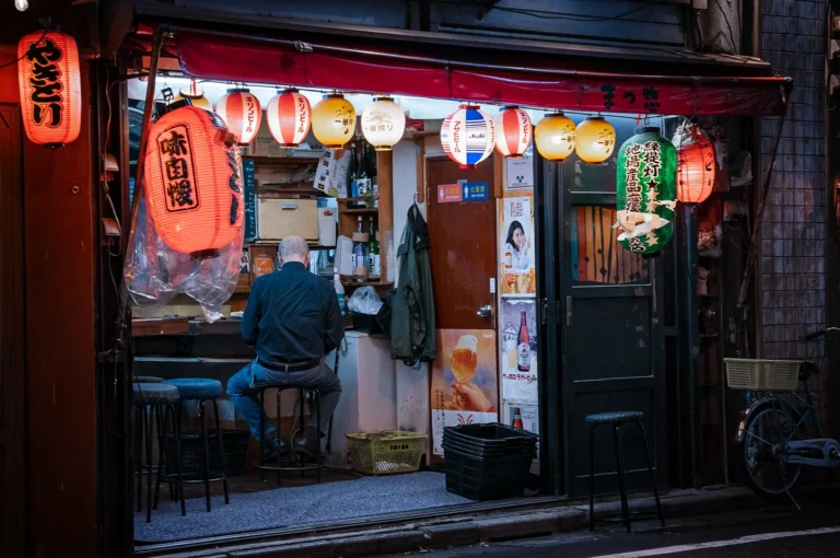 A wide, eye-level shot captures a quiet and intimate nighttime street scene at a traditional Japanese izakaya, representing the file "1-The Last Customer at the Counter Staying Until the End.webp". An elderly, bald man wearing a dark long-sleeve shirt and blue jeans sits on a low stool with his back to the camera, looking toward the interior counter of a small, open-front restaurant. The storefront is framed by a low red awning and illuminated by a bright, vibrant row of hanging paper lanterns in shades of red, white, yellow, and green adorned with Japanese text and logos. Vintage beverage posters and a green coat hang on the wall next to a sliding door, while empty stools, plastic crates, and a bicycle parked on the right emphasize the calm, end-of-the-night atmosphere of the alleyway.