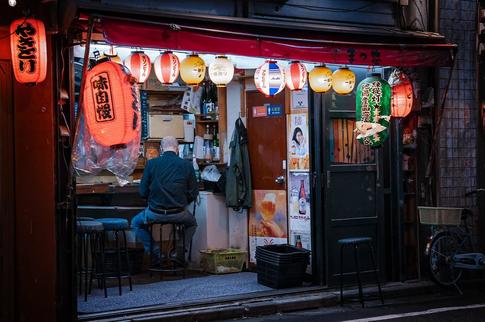 A wide, eye-level shot captures a quiet and intimate nighttime street scene at a traditional Japanese izakaya, representing the file "1-The Last Customer at the Counter Staying Until the End.webp". An elderly, bald man wearing a dark long-sleeve shirt and blue jeans sits on a low stool with his back to the camera, looking toward the interior counter of a small, open-front restaurant. The storefront is framed by a low red awning and illuminated by a bright, vibrant row of hanging paper lanterns in shades of red, white, yellow, and green adorned with Japanese text and logos. Vintage beverage posters and a green coat hang on the wall next to a sliding door, while empty stools, plastic crates, and a bicycle parked on the right emphasize the calm, end-of-the-night atmosphere of the alleyway.
