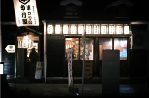 A wide, eye-level night shot captures the glowing storefront of a traditional Japanese izakaya or kushikatsu restaurant, representing the file "1-What Happens After the Last Order.webp". Standing out against the dark night, the restaurant features a dark wood facade illuminated by a row of ten bright white paper lanterns arranged horizontally above the main glass sliding doors. On the left, a smaller entryway with a white noren curtain and a single warm hanging bulb shows a silhouette of a person inside, while a large, angular illuminated sign extends outward showcasing bold Japanese calligraphy. In the foreground, a quiet pavement with a vertical banner and a street sign post completes the atmospheric, late-night dining scene.