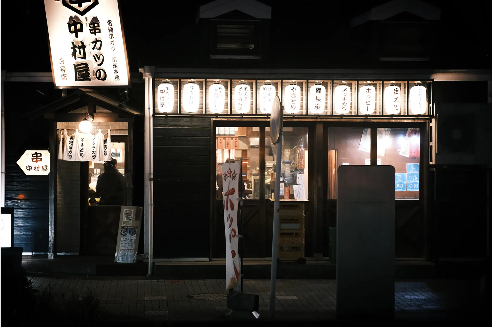 A wide, eye-level night shot captures the glowing storefront of a traditional Japanese izakaya or kushikatsu restaurant, representing the file "1-What Happens After the Last Order.webp". Standing out against the dark night, the restaurant features a dark wood facade illuminated by a row of ten bright white paper lanterns arranged horizontally above the main glass sliding doors. On the left, a smaller entryway with a white noren curtain and a single warm hanging bulb shows a silhouette of a person inside, while a large, angular illuminated sign extends outward showcasing bold Japanese calligraphy. In the foreground, a quiet pavement with a vertical banner and a street sign post completes the atmospheric, late-night dining scene.