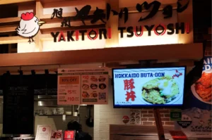 A medium, eye-level storefront shot captures the ordering counter of a Japanese food stall named "Yakitori Tsuyoshi," representing the file "1-Yakitori Tsuyoshi Review What_2.webp". A large wooden sign features the restaurant's name in bold black and red lettering alongside a cute cartoon chicken illustration and Japanese calligraphy. Below the sign, a digital menu screen on the right advertises a "Hokkaido Buta-Don" pork bowl dish, while printed promotional posters detailing a loyalty program and a chalkboard menu listing chicken wings hang to its left. Dark fabric noren curtains partially obscure the brightly lit kitchen area in the background, completing the casual counter-service dining setup.