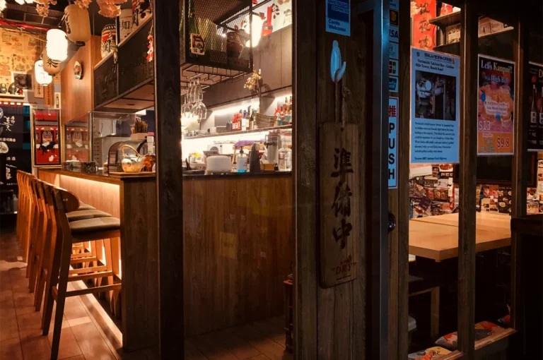 A wide, eye-level shot framed through a wooden doorway captures the warm, cozy entrance of an authentic Japanese restaurant, representing the file "1-Yorimichi Yakitori Izakaya Review.webp". On the left, a long wooden bar counter illuminated by a soft golden under-counter glow is lined with a row of empty, high-backed wooden barstools with dark cushions. Behind the bar, an organized kitchen and drink preparation station features rows of glasses and bottles under glowing paper lanterns hanging from the ceiling. To the right, a glass windowpane features a prominent vertical "PUSH" sign next to a hanging wooden plaque inscribed with Japanese characters, alongside promotional posters and reviews displayed for incoming diners.