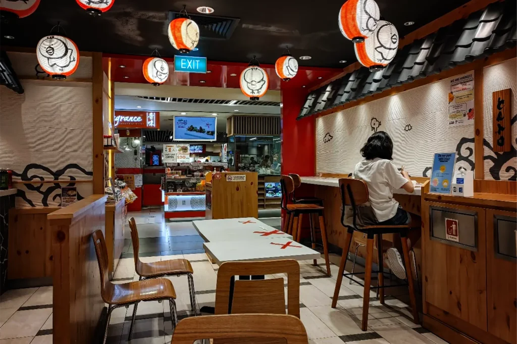 A wide, eye-level shot captures the brightly lit interior of a Japanese casual dining restaurant, representing the file "2-Best Yakitori Near Plaza Singapura.webp". On the right, a lone customer with short dark hair, wearing a white t-shirt, sits on a high wooden barstool at a long counter that is decorated with a faux tiled roof overhang and simple wave graphics on the wall. The dining area features several low wooden tables and chairs, with large red tape "X" marks placed across the center tables to indicate social distancing. Hanging from the dark ceiling are several round, glowing orange and white paper lanterns adorned with stylized chicken illustrations, while the open background reveals a brightly lit mall corridor with a green illuminated "EXIT" sign and a glimpse of a food kiosk labeled "Fragrance."